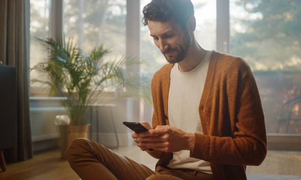 Man Using Smartphone at Home, Does Remote Work. Handsome Smiling Male Sitting on the Floor Works with Papers, Documents, Brainstorms Creative Project with Online Research. Sunny Living Room
